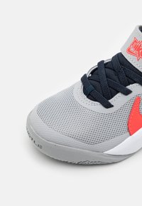 Harmaa urheilukenkä, jossa on teksturoitu verkkomateriaali, tummansinisiä nauhoja ja punainen yksityiskohta. Ominaisuuksiin kuuluu kumiulkopohja ja vahvistettu varvasosa.