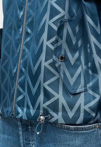Veste bleue avec motif MK répétitif, fermeture éclair argentée, poche à rabat avec bouton de marque et cordon de serrage ajustable à la taille, portée sur un jean bleu.