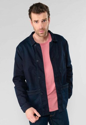 Veste à boutons navy avec col, deux poches avant, portée sur une chemise rose, associée avec un jean foncé. Texture lisse et design décontracté.