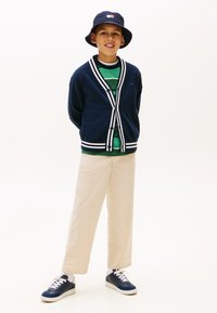 Αγόρι που φοράει ναυτικό καπέλο τύπου μπαουκ, ναυτικό κάρντιγκαν με λευκές ρίγες, ριγέ πράσινο πουκάμισο, μπεζ παντελόνι και ναυτικά αθλητικά παπούτσια με λευκές σόλες, στέκεται και χαμογελά.