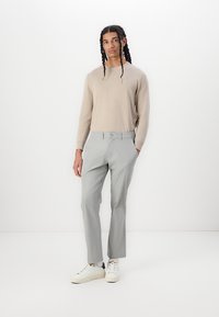 Lys beige sweater, tætsiddende; lysegrå bukser med en glat tekstur; hvide sneakers; stående stilling mod en ensfarvet baggrund.