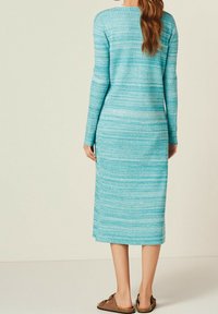 Femme se tenant de dos, portant une robe bleue texturée à manches longues et mi-mollet, ainsi que des sandales brunes à enfiler, sur un fond uni.