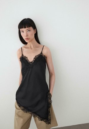 Femme aux longs cheveux foncés portant une camisole noire bordée de dentelle sur un pantalon beige, debout contre un mur clair uni, mains dans les poches.