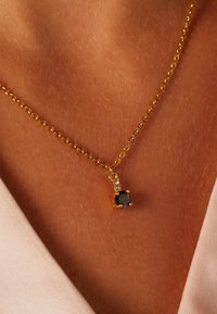 Collier en chaîne dorée arborant un pendentif en pierre noire avec des accents clairs. Le collier repose sur une surface couleur peau, mettant en valeur des maillons détaillés.
