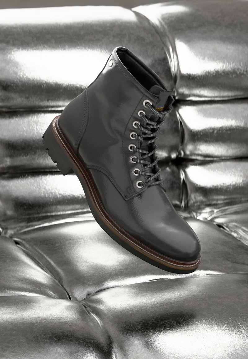 Sorte lær ankle boots med en glat finish, sølvøjer og grå snørebånd, hvilende på en skinnende sølv tufted overflade.