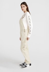 Cream-colored overalls with adjustable straps, side pockets, and a logo accent. Paired with a long-sleeve white top featuring black patterns.