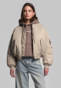 Beige bomberjakke lavet af glat materiale, med ribbede ærmer, to sidelommer og en lynlåslomme på ærmet. Bæres over en brun hoodie.