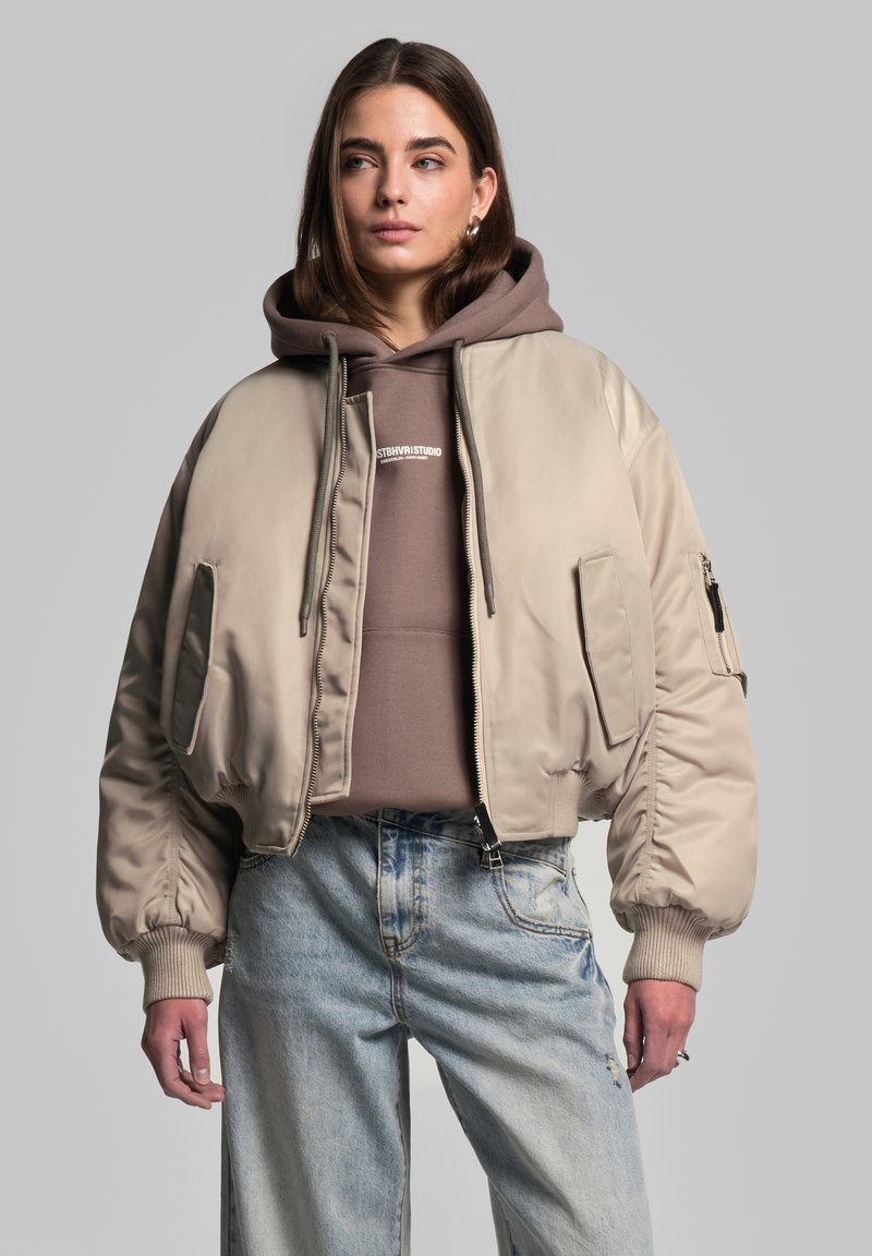 Beige bomberjakke lavet af glat materiale, med ribbede ærmer, to sidelommer og en lynlåslomme på ærmet. Bæres over en brun hoodie.