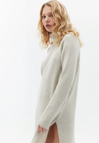 Vestido de punto en gris claro con textura de canalé vertical, cuello alto y abertura lateral, con mangas largas y un corte relajado.