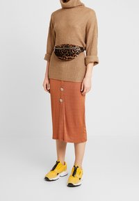Brązowy sweter z golfem, rdzawe midi spódnica w prążki z guzikami, czarna torba na pasek w panterkę oraz żółte sneakersy z czarnymi akcentami.