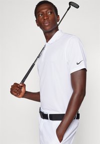 Biela polokošeľa s krátkymi rukávmi, čierne logo Nike na rukáve, skombinovaná s bielymi nohavicami a čiernym opaskom, drží golfovú palicu na ramene.