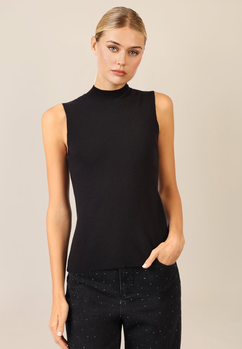 Top senza maniche nero realizzato in maglia con collo alto; texture liscia e design aderente. Indossato con jeans scuri caratterizzati da sottili decorazioni.