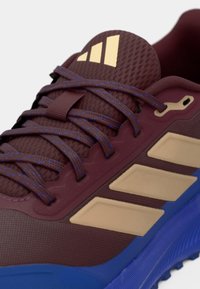 Viininpunainen lenkkikenkä teksturoidulla päällisellä, sinisillä yksityiskohdilla ja kultaisella logolla. Varustettu litteillä nauhoilla ja kuvioidulla pohjalla pitoon.