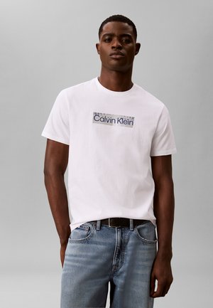 Muž v bielom tričku s logom Calvin Klein a modrých džínsoch stojaci pred jednofarebným svetlosivým pozadím.