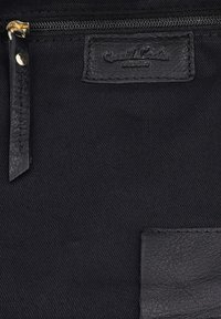 Schwarze Stoffoberfläche mit einem Reißverschluss-Zieher aus Leder und zwei aufgenähten schwarzen Lederetiketten, von denen eines mit dem Text "Guest Roles studio" geprägt ist.