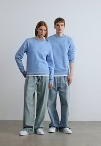 Ανοιχτό μπλε φούτερ με ριμπ μανσέτες, φορεμένα πάνω από λευκά T-shirts. Συνδυασμένα με φαρδιά ξεβαμμένα τζιν και λευκά αθλητικά παπούτσια. Ελάχιστη ορατή εμπορική σήμανση.