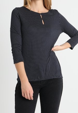 Marineblauw polkadot top met driekwart mouwen en een keyhole-uitsparing, gemaakt van soepele stof, aangesloten silhouet, gecombineerd met donkere jeans.