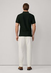 Muž stojící zády, oblečený v černém polo tričku s krátkým rukávem, bílých kalhotách a hnědých botách, na jednobarevném pozadí.