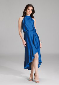 Niebieska sukienka na ramiączkach w kolorze satyny z dopasowanym gorsetem i falbaniastym asymetrycznym dołem. Połączona z srebrnymi sandałami na obcasie. Gładka tekstura.