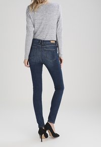 Mujer vestida con jeans ajustados de color azul oscuro, blusa de manga larga gris y tacones altos negros, vista desde atrás contra un fondo liso.