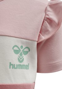 Pink bomulds T-shirt med rynkede ærmer, der har et hvidt panel og et lys grønt "hummel" logo med et stiliseret emblem ovenfor.
