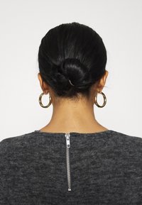 Maglione grigio con finitura testurizzata, dotato di una cerniera metallica visibile sullo scollo posteriore e orecchini a cerchio dorati. Capelli raccolti in uno chignon.