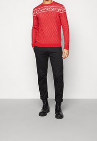 Maglione rosso lavorato a maglia con motivi geometrici e renne bianchi, abbinato a pantaloni neri e stivali neri con lacci.