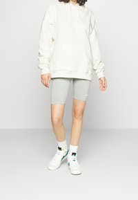 Sudadera blanca con capucha, gran bolsillo frontal y cordones, combinada con pantalones cortos de ciclismo grises y zapatillas blancas con detalles verdes. Se aprecian texturas suaves.