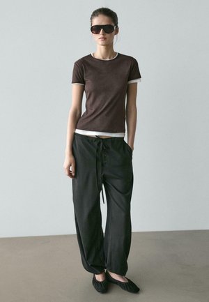 Femme debout, portant des lunettes de soleil foncées, un t-shirt marron ajusté sur un sous-t-shirt blanc, un pantalon noir ample avec cordon de serrage, et des ballerines noires.