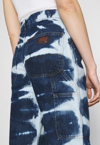 Jeanscargo-byxor med ett blått och vitt tie-dye-mönster, bakficka med märkesetikett, synliga sömmar och en avslappnad passform.
