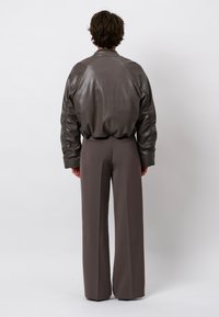 Persoon die met de rug naar de kijker staat, gekleed in een losse donkergrijze leren jas en brede donkere pantalons tegen een effen achtergrond.