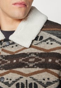 Wollen blend overhemdjack met een witte nepbont kraag. Bevat abstracte patronen in bruin, crème en zwart. Inclusief knoopsluiting.