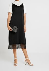 Černé šaty s krajkovým lemem, nošené přes bílé tričko s krátkým rukávem. Bílá sandály na podpatku a černé psaníčko doplňují outfit.