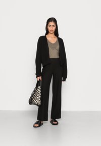 Cardigã preto canelado sobre um top bege com decote adornado, combinado com calças largas pretas e sandálias de slide pretas, segurando uma bolsa texturizada.