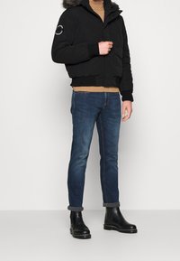 Man wearing black hooded jacket with fur trim, tan sweater, blue rolled-up jeans, and black leather boots standing against white background.