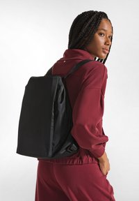 Zaino in tessuto nero con design rettangolare, spallacci regolabili, superficie liscia e dettaglio logo discreto sulla parte anteriore.