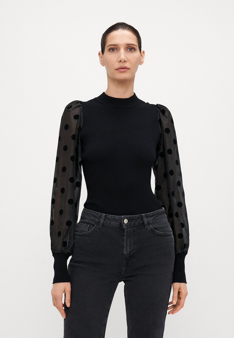 Svart ribbad långärm topp med genomskinliga, puffiga prickiga ärmar; kombinerad med mörka jeans; minimalistisk design och åtsittande silhuett.