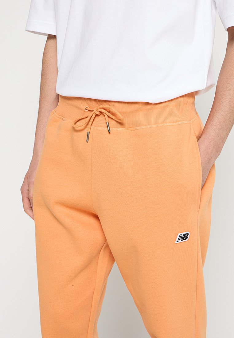 Pantalones de chándal naranjas con cintura ajustable, con un pequeño logo negro en el muslo izquierdo. Textura de tela suave y cinturilla acanalada.