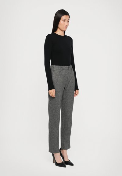 Schwarzes Langarmoberteil kombiniert mit grauen, gemusterten Hosen und schwarzen spitzen Absätzen; das Outfit weist eine taillierte Silhouette und einen elastischen Taillenbund auf.