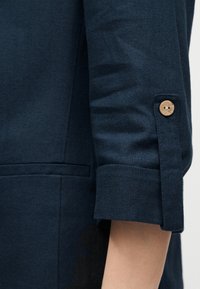 Manche d'un vêtement bleu foncé remontée jusqu'au coude, maintenue par un bouton en bois sur une bride en tissu.