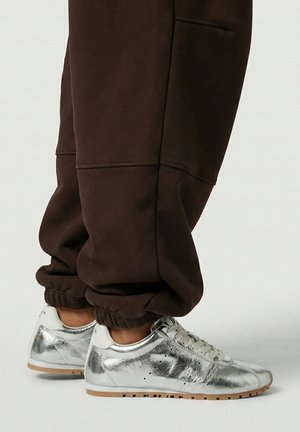 Pieds chaussés de baskets argentées métalliques brillantes associées à un pantalon de survêtement marron foncé retroussé, sur un fond uni.