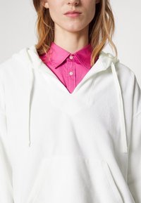 Bílá mikina s kapucí a jemnou texturou, s tkaničkami a přední kapsou, vrstvená přes jasně růžovou košili s límečkem.