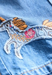 Tissu en denim avec patch cheval brodé présentant une crinière orange, une fleur rose et des coutures décoratives en fils bleu, brun et blanc.