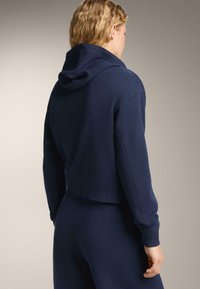 Krátka námornícky modrá mikina s pletenou textúrou, s kapucňou a priliehavými rukávmi. Tkanina sa zdá byť mäkká a tepl�á, určená na bežné nosenie.