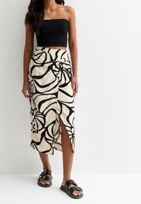 Zwarte strapless top gecombineerd met een beige rok met hoge taille, voorzien van grote zwarte bloemenpatronen en een split aan de zijkant. Zwarte sandalen met gespen.