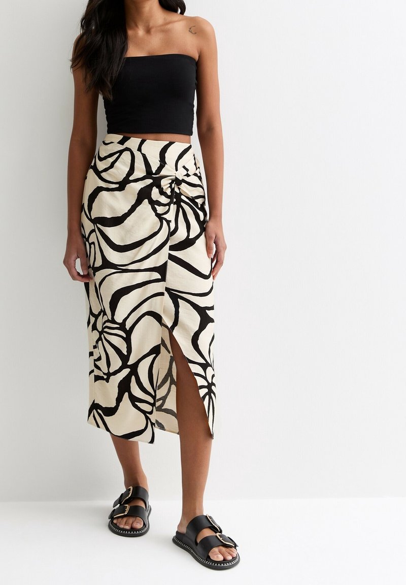 Zwarte strapless top gecombineerd met een beige rok met hoge taille, voorzien van grote zwarte bloemenpatronen en een split aan de zijkant. Zwarte sandalen met gespen.
