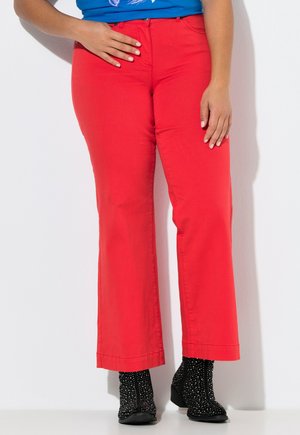 Nainen, jolla on kirkkaanpunaiset housut, mustat nilkkurit valkoisilla pisteillä ja sininen paita, seisoo yhdellä kädellä vyötäröllä ja toinen rentona.