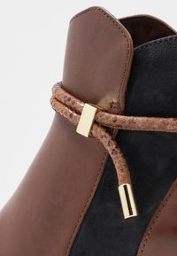 Bottine en cuir marron avec accents en suède noir, dotée d'une lanière décorative marron et or et d'une finition lisse et élégante.