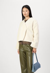Elfenben fuskraftjakke med en cropped design, kombinert med en brun leopardmønstret topp og grønne skinnbukser; holder en lyseblå håndveske.