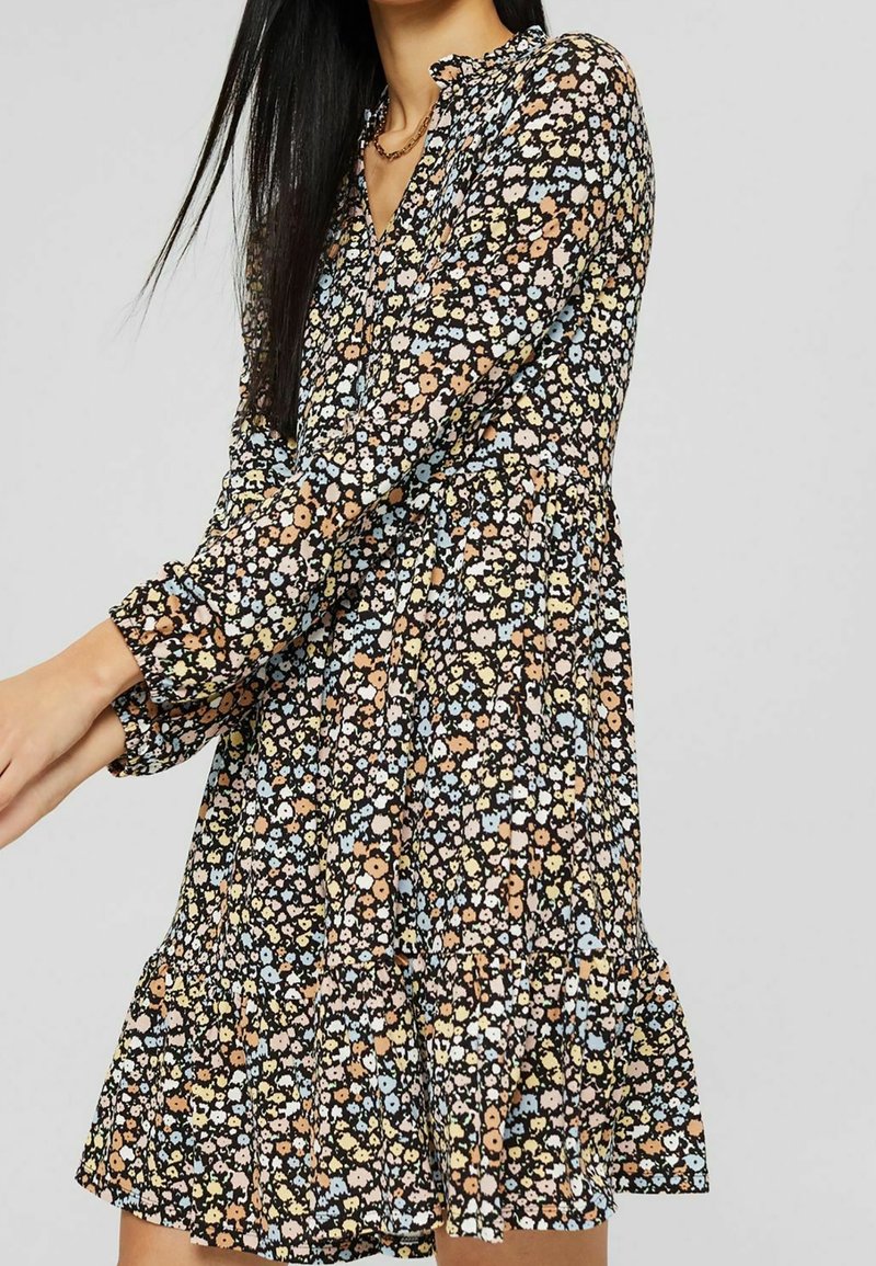 Robe florale noire avec une jupe à volants, des manches longues et un décolleté en V. Le tissu présente de petites fleurs colorées dans des nuances de bleu, pêche et jaune.